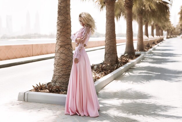 Bionda formosa vicino al mare a Dubai, palme, abiti caldi e splendidi, servizio fotografico estivo di stile di vita soleggiato, sventolando il vestito dal vento, calma e relax vicino alla piscina, acconciatura, trucco