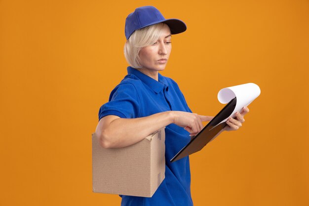 Bionda premurosa donna delle consegne in uniforme blu e cappuccio in piedi in vista di profilo tenendo la scatola di cartone sotto le ascelle e appunti guardando gli appunti mettendo il dito su di esso