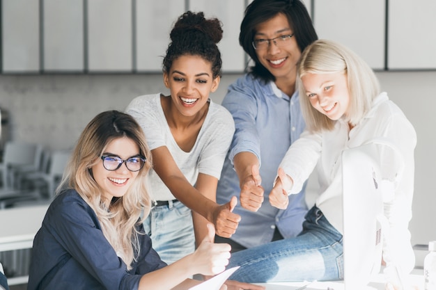 Bionda specialista in informatica che scherza con gli amici, seduta sul posto di lavoro e ride. Responsabile giapponese emozionante che posa con il sorriso, in piedi accanto al collega biondo in ufficio.