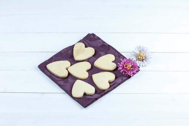 Biscotti a forma di cuore di vista di alto angolo in tovaglietta con i fiori sul fondo del bordo di legno bianco. orizzontale