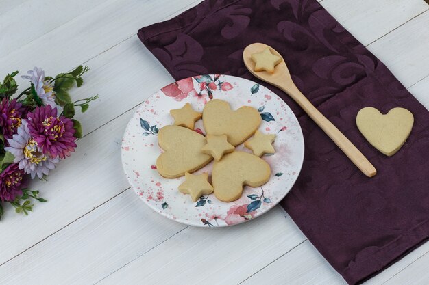 Biscotti in piatto e cucchiaio di legno con vista di alto angolo di fiori su fondo in legno e tessile