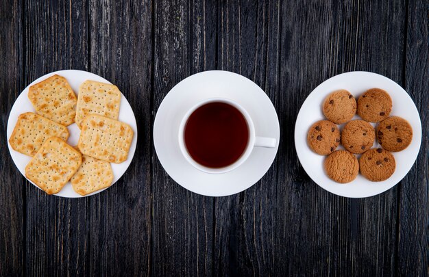 Biscotti salati del cioccolato dei cracker degli spuntini di vista superiore e tazza di tè su fondo di legno nero