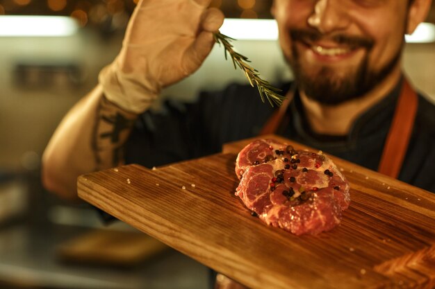Bistecca di manzo saporita con sale e pepe sul tagliere di legno chef che decora la carne con rosmarino uomo sorridente con la faccia barbuta in guanti e tatuaggio sul braccio sullo sfondo