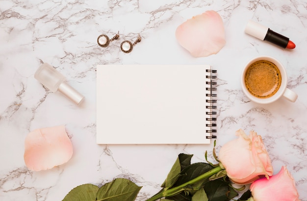 Blocco note a spirale in bianco con orecchini; bottiglia di smalto per unghie; rossetto; rose e tazza di caffè su priorità bassa di marmo