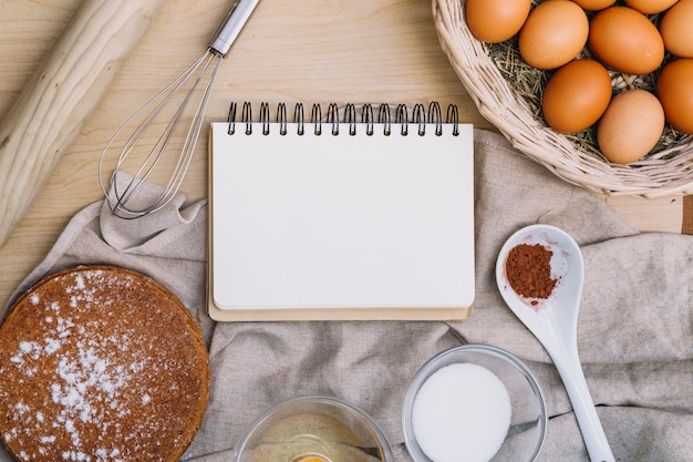 Blocco note a spirale in bianco con vari ingredienti del dolce di cottura sulla tavola