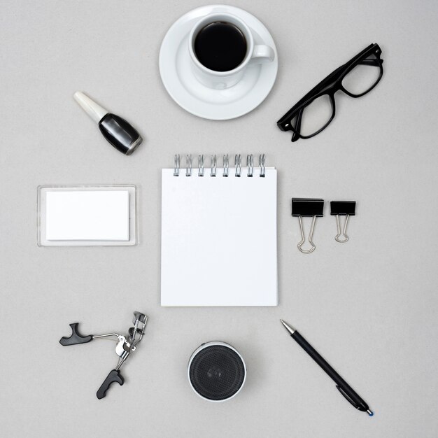 Blocco note bianco vuoto circondato da una tazza di caffè; smalto; bigodino per ciglia; altoparlante; penna e graffette sopra fondo grigio