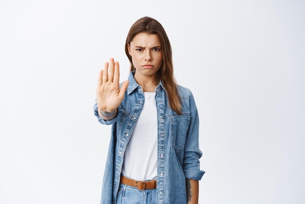 Blocco Una donna seria che dice di non accigliarsi e di estendere la mano per il gesto di arresto vieta qualcosa di deludente in piedi su sfondo bianco