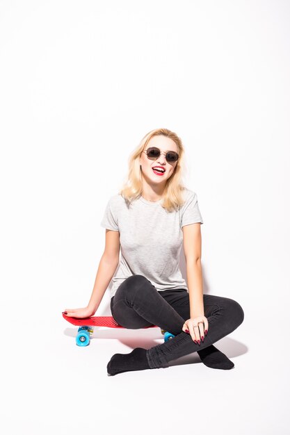 Blondie con le gambe incrociate si siede su skateboard rosso davanti al muro bianco