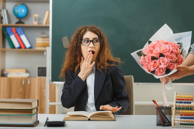 bocca coperta sorpresa con la mano la giovane insegnante femminile ha ricevuto un mazzo di fiori seduto alla scrivania con gli strumenti della scuola in classe