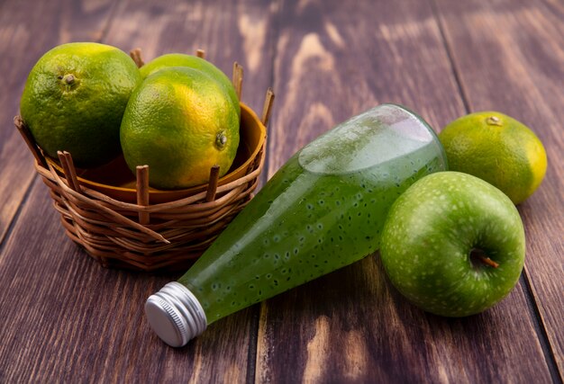 Bottiglia del succo di vista frontale con la merce nel cestino dei mandarini e la mela sulla parete di legno