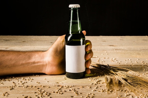 Bottiglia di birra della tenuta della mano dell&#39;uomo con le orecchie di grano su superficie di legno