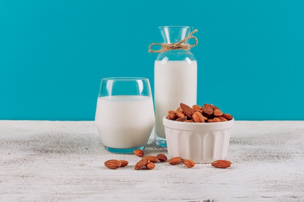 Bottiglia di latte con bicchiere di latte e ciotola di mandorle vista laterale su uno sfondo bianco in legno e blu