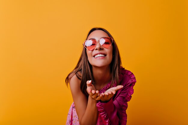 Brillante di felicità, la giovane ragazza maliziosa con gli occhiali da sole rosa sta mandando un bacio d'aria. Chiudere il ritratto di carino donna dai capelli castani