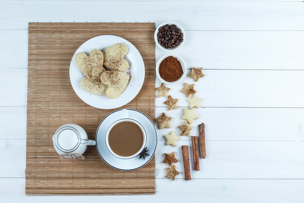 Brocca di latte, tazza di caffè, biscotti a forma di cuore su una tovaglietta con chicchi di caffè e farina, biscotti a stella, cannella vista dall'alto su un fondo di tavola di legno bianco