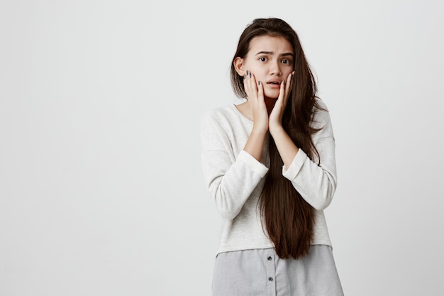 Bruna donna adolescente con gli occhi scuri sollevando le sopracciglia, fissando con grandi occhi, tenendosi per mano sulle guance, essendo nel panico preoccupata di sentire cattive notizie. Giovane femmina inorridita con sguardo spaventato scioccato