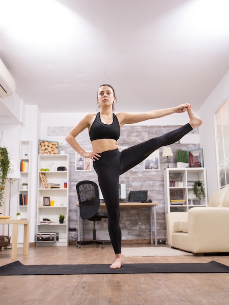 Bruna donna adulta che fa una posa di yoga gamba in piedi nel soggiorno.