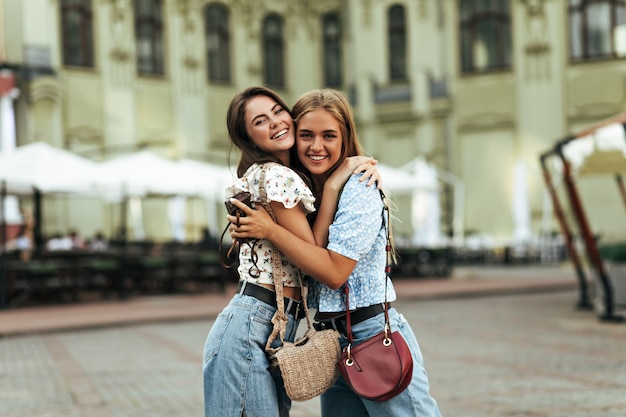 Bruna gioiosa e giovane donna bionda in eleganti pantaloni di jeans e camicette colorate si rallegrano, si divertono, sorridono ampiamente all'aperto