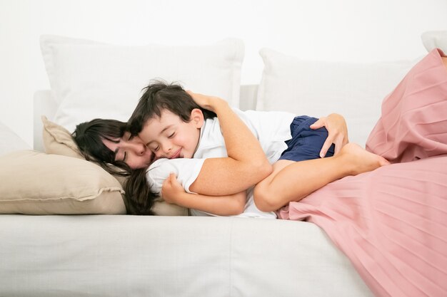 Bruna madre che dorme sul divano e abbraccia il figlio carino.