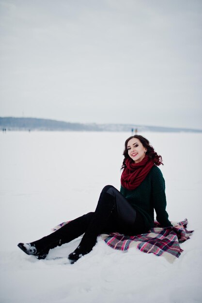 Bruna ragazza in maglione verde e sciarpa rossa seduta sul plaid lago ghiacciato all'aperto la sera il giorno d'inverno