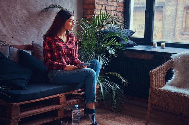 Brunette affascinante in una camicia di flanella e jeans che si siede su un couc