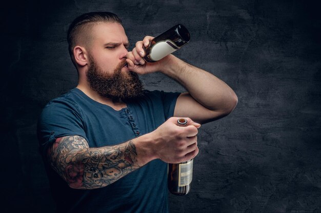 Brutale maschio barbuto con braccio tatuato beve una birra da una bottiglia.