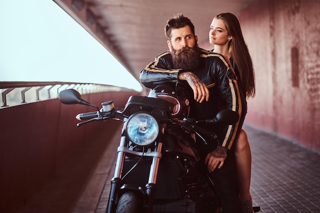 Brutale motociclista barbuto in giacca di pelle nera e sensuale ragazza bruna seduta insieme su una moto retrò su misura su un marciapiede sotto un ponte.