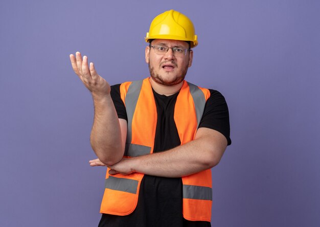 Builder uomo in costruzione giubbotto e casco di sicurezza guardando la telecamera confuso e dispiaciuto con il braccio fuori come sostenendo in piedi sul blu