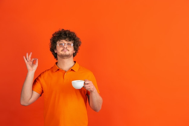 Buon caffè, felice. Ritratto monocromatico del giovane caucasico sulla parete arancione. Bellissimo modello riccio maschio in stile casual.