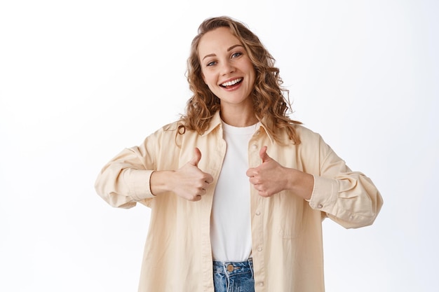 Buon lavoro. La donna bionda sorridente mostra i pollici in su in segno di approvazione, dicendo sì, d'accordo e come il prodotto, loda la scelta, in piedi su sfondo bianco