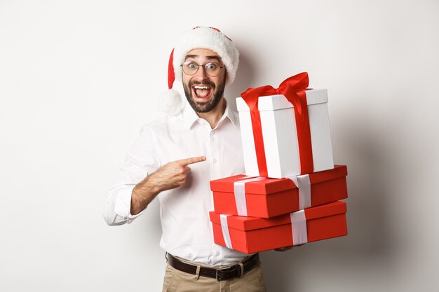 Buon Natale, concetto di vacanze. L'uomo sorpreso riceve i regali di natale, indicando i regali e sorridendo felice, portando il cappello della Santa
