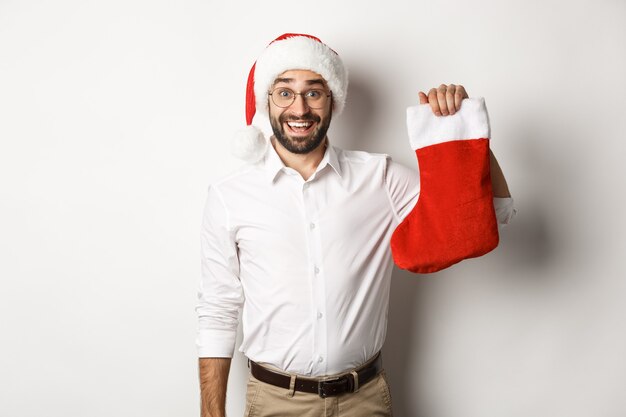 Buon Natale, concetto di vacanze. Ragazzo barbuto eccitato in cappello della santa che tiene il calzino di Natale e sorridente, che celebra il nuovo anno