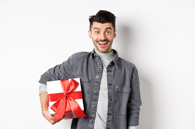 Buon San Valentino. Ragazzo sorpreso e felice che tiene in mano una scatola regalo e guarda la macchina fotografica, sorride stupito, celebra le vacanze, porta un regalo ad un appuntamento romantico, sfondo bianco