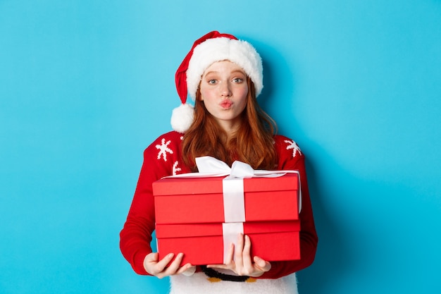 Buone vacanze e concetto di Natale. Carina ragazza rossa con regali e labbra arricciate per un bacio, indossando un cappello da Babbo Natale e un maglione divertente, sfondo blu.