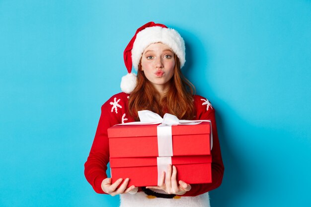 Buone vacanze e concetto di Natale. Carina ragazza rossa con regali e labbra arricciate per un bacio, indossando un cappello da Babbo Natale e un maglione divertente, sfondo blu.