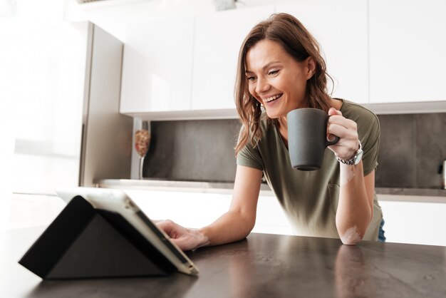 Caffè bevente della donna casuale felice e per mezzo del computer della compressa sulla cucina