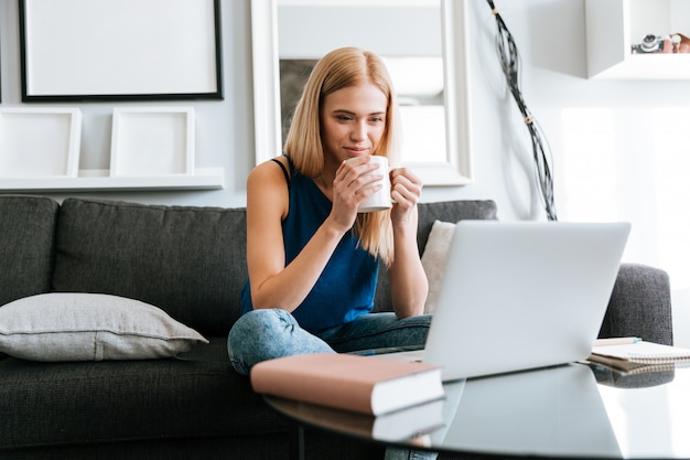 Caffè bevente della donna premurosa e computer portatile usando a casa