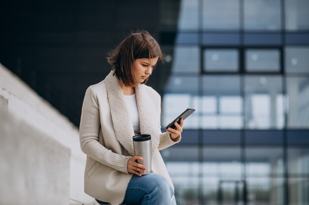 Caffè bevente della giovane donna e telefono usando usando fuori della via