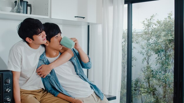 Caffè bevente delle coppie gay asiatiche, divertendosi a casa. I giovani uomini LGBTQ + che parlano felici si rilassano insieme riposano insieme il tempo romantico nella cucina moderna a casa la mattina.