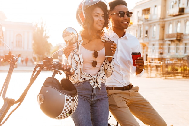 Caffè bevente delle giovani coppie africane felici mentre stando vicino alla motocicletta moderna sulla via e sul distogliere lo sguardo