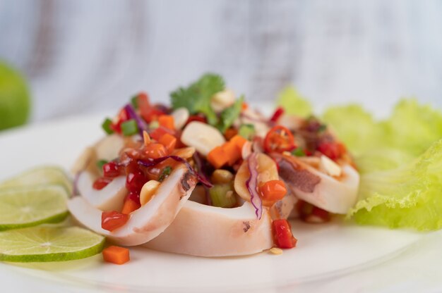 Calamaro piccante al limone con galanga, peperoncini rossi, carote, menta piperita, cipollotti e aglio su un piatto su un pavimento di legno bianco.