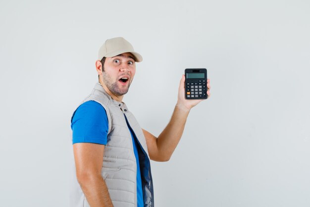 Calcolatrice della holding del giovane in maglietta, giacca e sguardo stupito.