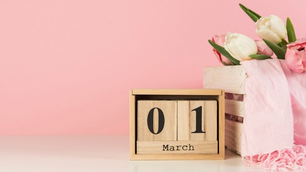 Calendario in legno con 1 marzo vicino alla cassa con tulipani e sciarpa sulla scrivania su sfondo rosa