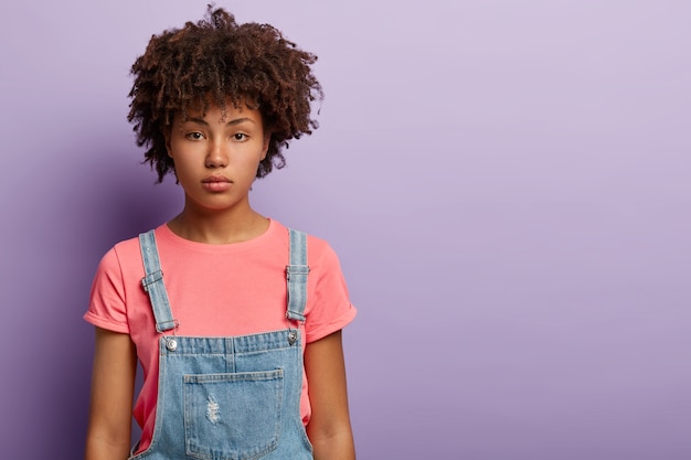 Calma attraente ragazza millenaria con taglio di capelli afro, pelle scura, senza trucco, vestita con una maglietta e una tuta di jeans, guarda seriamente la telecamera