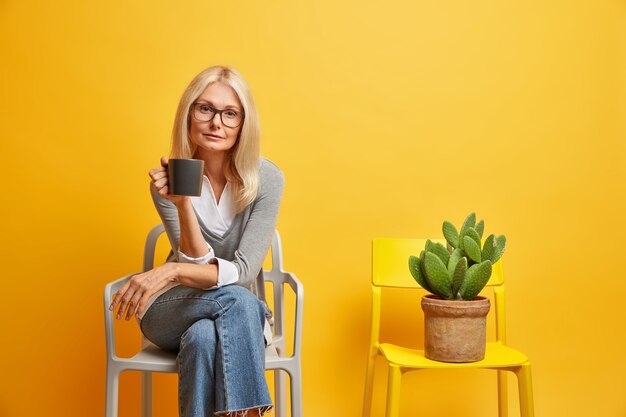 calma donna bionda si siede su una comoda sedia con bevanda guarda con sicurezza e posa vicino a cactus in vaso gode di un'atmosfera tranquilla. Concetto di stile di vita