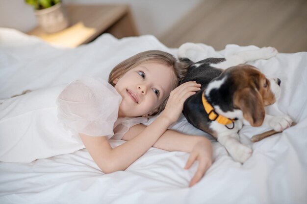 Calma e carina bambina caucasica dai capelli lunghi sdraiata sul letto accanto al suo amico a quattro zampe