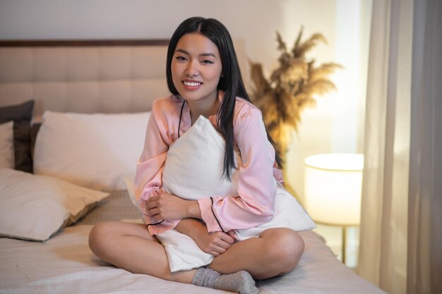 Calma. Giovane donna asiatica sorridente adulta con lunghi capelli scuri in pigiama rosa seduto sul letto che abbraccia cuscino guardando la fotocamera