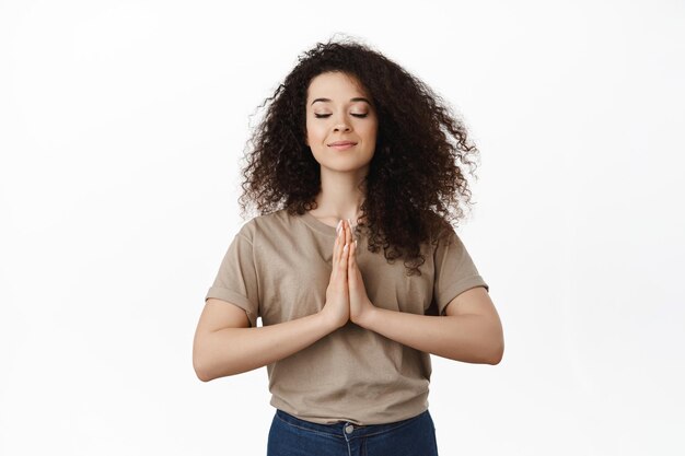 Calma giovane donna meditando, mostra namaste pregare gesto, chiudere gli occhi e sorridere, esprimere desideri, supplicare Dio, in piedi su sfondo bianco