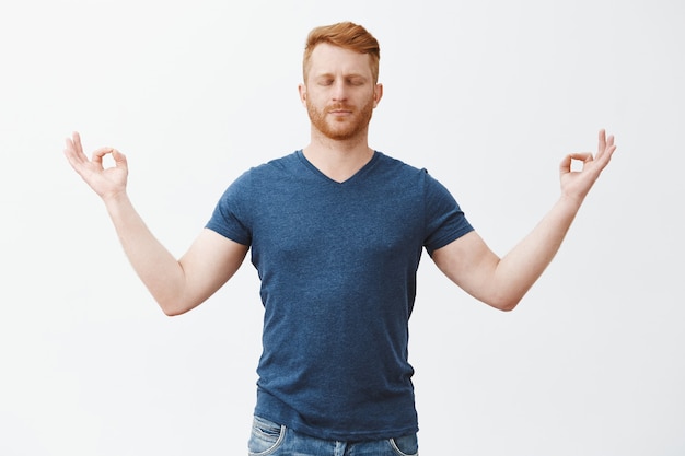 Calmati, pace dentro. Ragazzo europeo rilassato di bell'aspetto con capelli rossi e setole, occhi chiusi, in piedi con le mani divaricate con gesto zen, meditazione, alleviare lo stress con lo yoga