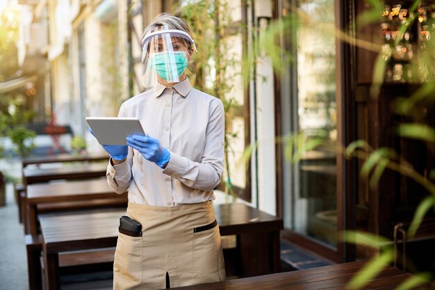 Cameriera che lavora sul touchpad mentre indossa una maschera protettiva e una visiera al bar all'aperto