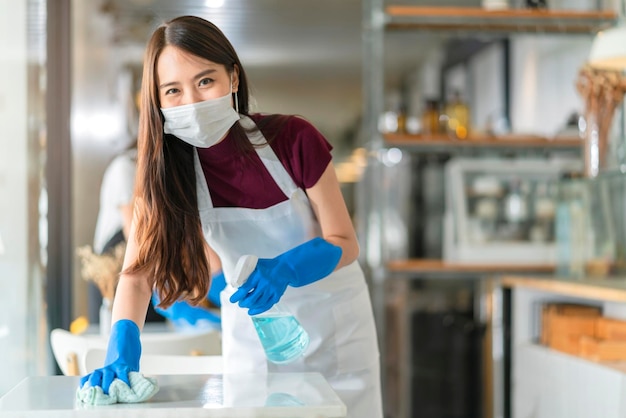 Cameriera del personale asiatico che indossa grembiule con maschera protettiva e tavolo per la pulizia dei guanti di gomma con spray alcolico disinfettante per prevenire l'infezione da coronavirus nella caffetteria bar nuovo concetto normale